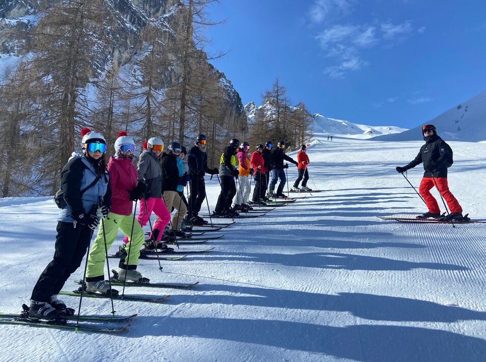 Eine Gruppe Jugendlicher steht mit Skiern in einer Reihe auf einer Piste, eine Person fährt vorbei
